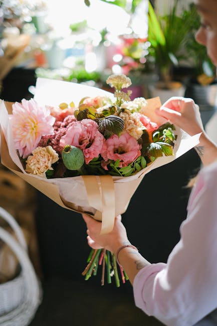 A person holding a large bouquet of fresh flowers, including pink peonies, cream-colored carnations, and white accents, wrapped in cream and white paper with a beige ribbon. The arrangement features l