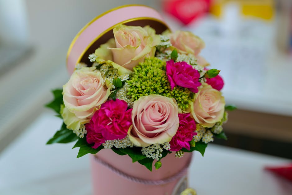 A close-up of a floral gift arrangement in a pastel pink round box, featuring soft pink roses, vibrant pink carnations, green hypericum berries, white baby's breath, and lush green leaves, arranged in