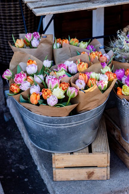 A galvanized metal tub filled with multiple bouquets of fresh tulips and ranunculus flowers wrapped in brown kraft paper, displaying a mix of pink, purple, orange, and yellow hues. The flowers have vi