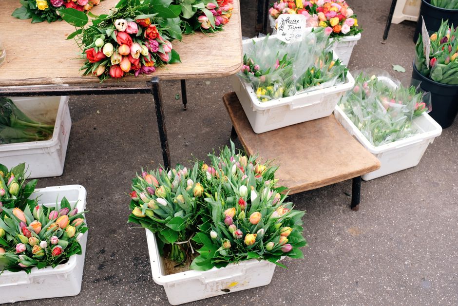 A collection of fresh tulip bouquets with pink, yellow, and white blooms and green leaves, arranged in white plastic crates and placed on rustic wooden tables outdoors. The tulips have smooth, slightl