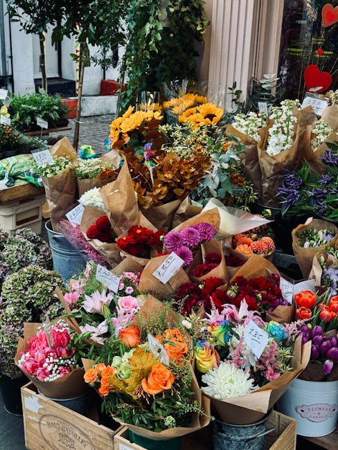 A vibrant display of freshly cut flowers arranged in brown kraft paper bouquets and metal buckets at a flower shop. The selection includes bright yellow sunflowers, soft pink lilies, deep red roses, p