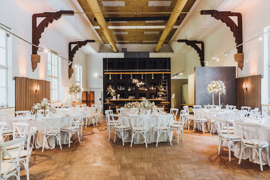 A spacious wedding reception venue with high ceilings and large windows allowing natural light to illuminate the room. The room features elegant round tables covered with white tablecloths, each toppe