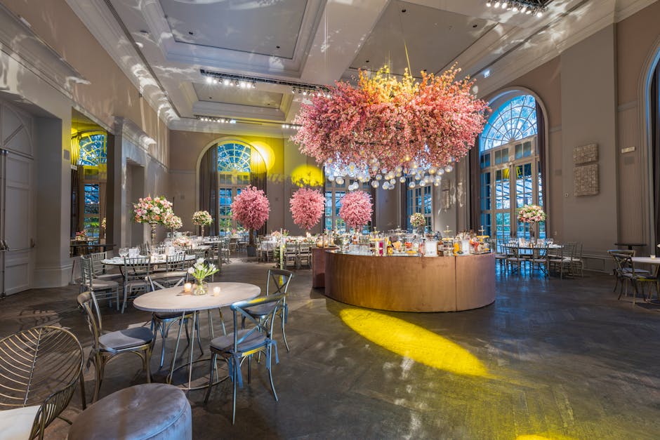 An elegant event hall decorated with an extensive floral installation featuring a large, spherical chandelier made of pink cherry blossoms and small white hanging orbs. Surrounding the centerpiece are