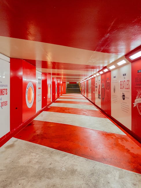 The image depicts a vibrant red underground walkway with a white and red patterned floor. The walls are decorated with various posters and signage in matching red hues, creating a lively atmosphere su